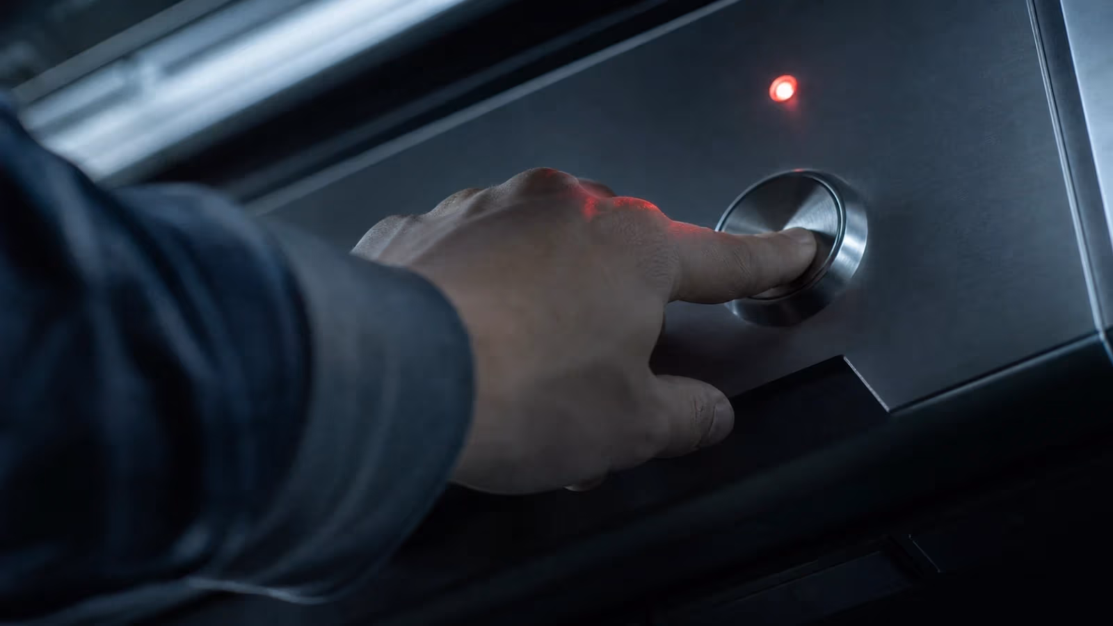 "Mano che preme un pulsante metallico su console industriale con una spia rossa accesa sopra"